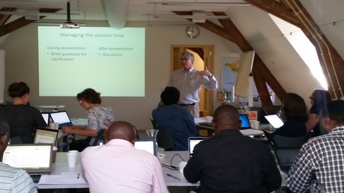 A standing European man speaking to a room of seated African and European men and women