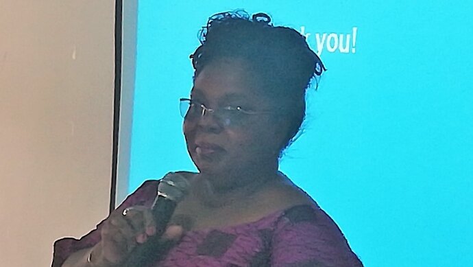 An African lady wearing a pink top and glasses speaks into a microphone