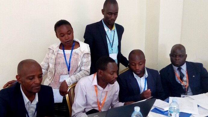 Six seated and standing African men and women, all looking and a laptop on a desk