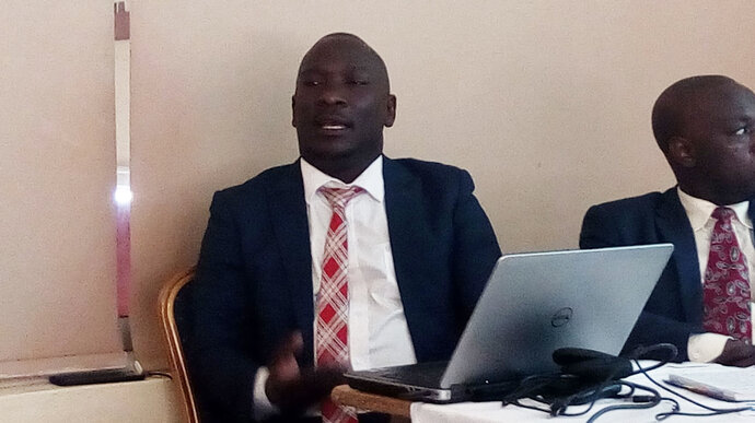 Seated African man in a suit speaking. He has a laptop open in front of him.