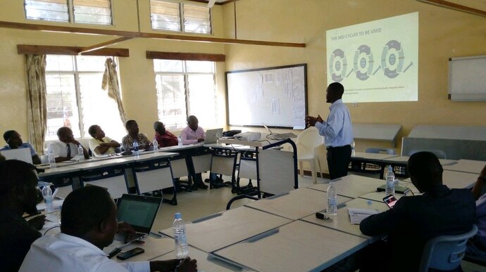 Man standing up in front of a projector screen while talking to people seated around him