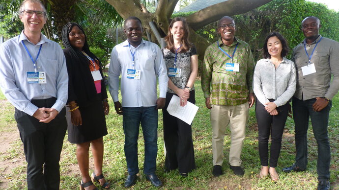 A row of seven standing, smiling African and European men and women in a garden