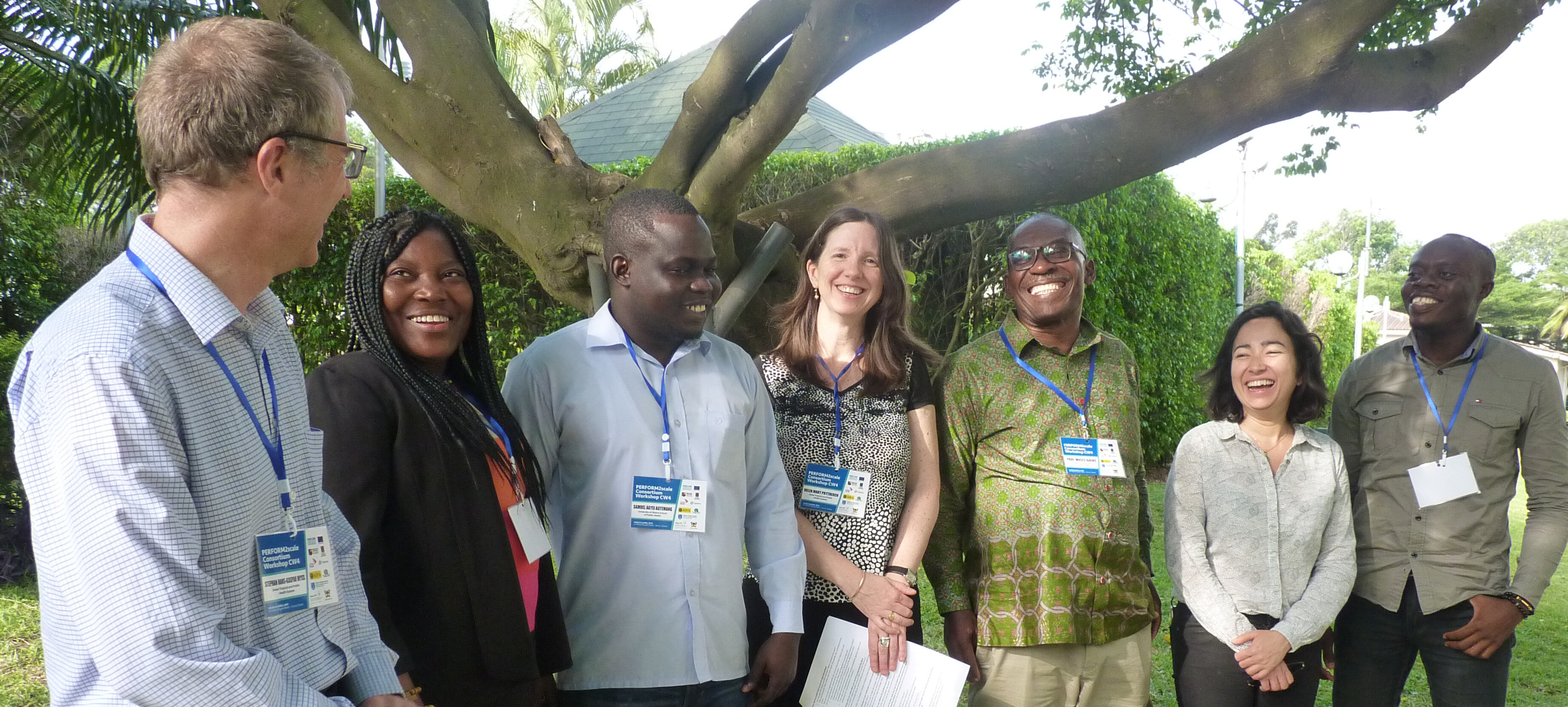 Seven smiling men and women, standing in front of a tree