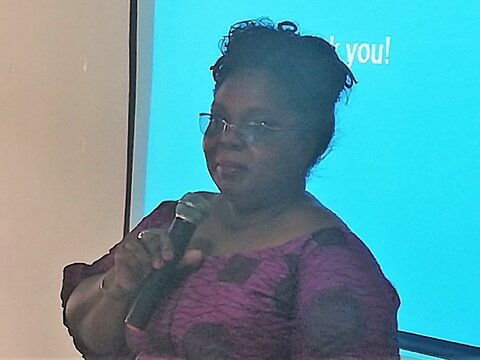 An African lady with a pink top and glasses speaks into a microphone