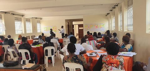 A group of about 30 African men and women, seated on white chairs while watchign man at the fron tof the room talking about a poster covered in post-its