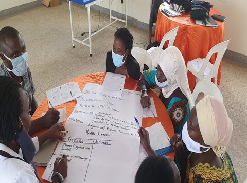 A group of masked African men and women sit around a table, writing on a piece of flipchart paper