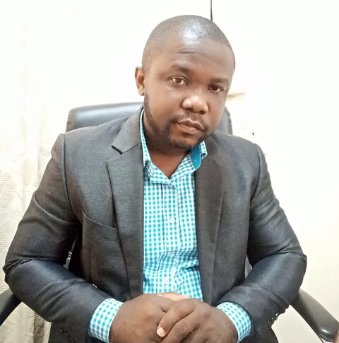 A sitting African man wearing a shirt and jacket looks at the camera